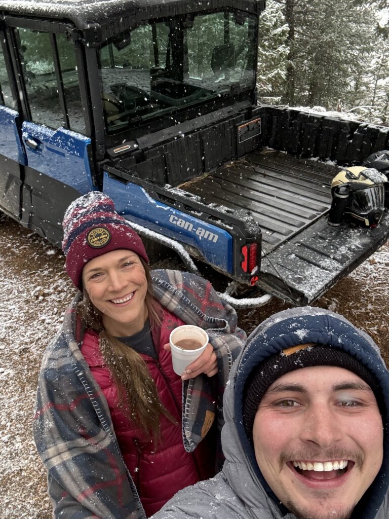 a selfie of Sarah the owner with a man drinking hot chocolate in the snow with a UTV in the background
